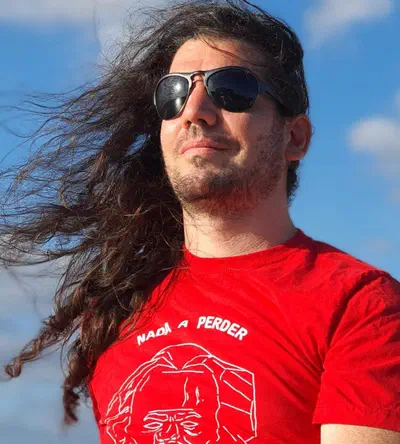 Headshot of Giovanni Bassi. He is white with long hair, is looking to the left, has his hair down, sunglasses and a red t-shirt, which reads 'nothing to lose', in Brazilian.